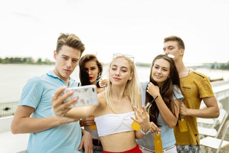 A company of good-looking friends laughing and drinking yellow cocktails and socialising and making selfie in the nice cafe next to the river. Cheers. Entertainment, having good time. River is in the background.の写真素材