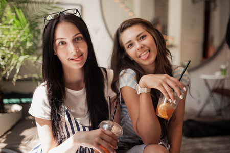 Two youthful smiling beautiful slim girls with long dark hair,wearing casual clothes, sit next to each other and look at the camera in a modern coffee shop.の写真素材