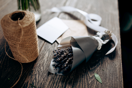 A little bouquet of flowers packed in dark paper, postcard, envelope, bobbin of twine and white ribbon are on the wooden tableの写真素材