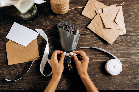 Florist ties a ribbon on the bouquet of flowers packed in dark paper on the wooden table with postcard, envelopes and  white ribbon on itの写真素材