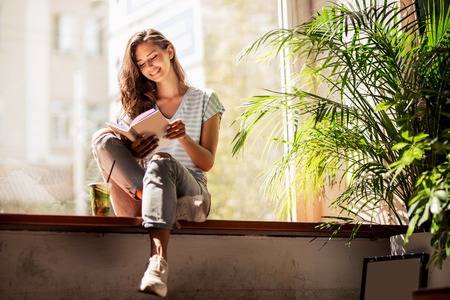 A pretty slim young girl with long hair,wearing casual outfit,sit on the windowsill  and reads a...