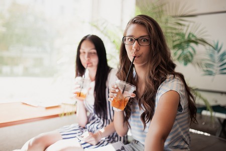 Two youthful smiling beautiful slim girls with long dark hair,wearing casual clothes, sit next to each other and look at the camera in a modern coffee shop.の写真素材