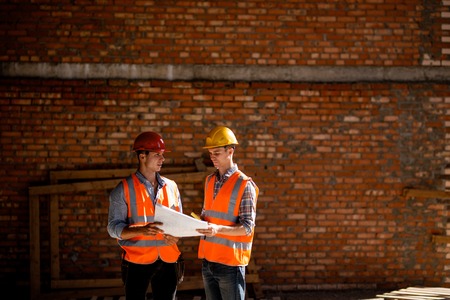 Architect  and construction manager dressed in orange work vests and helmets discuss documentation...