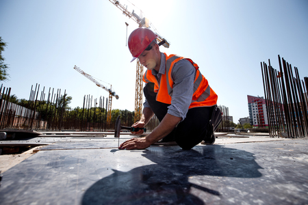 Builder dressed in orange work vest and helmet hammers a nail on the open building siteの写真素材