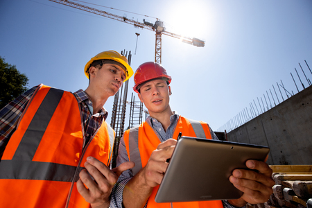 Architect  and structural engineer dressed in orange work vests and  helmets discuss a building...