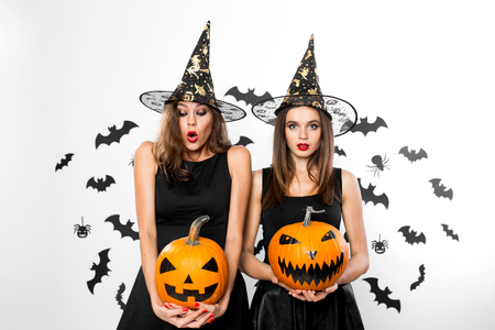 Two brunette girls in black dresses and witch`s hats hold horror halloween pumpkins on the background with batsの写真素材