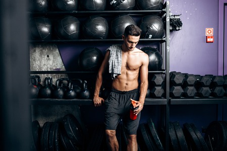 Athletic man with a naked torso and t-shirt on his shoulder dressed in the  black shorts stands near the sport equipment in the gymの写真素材