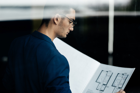 Architect dressed in stylish clothes stands next to window and explores the sheet with drawing in his handsの写真素材