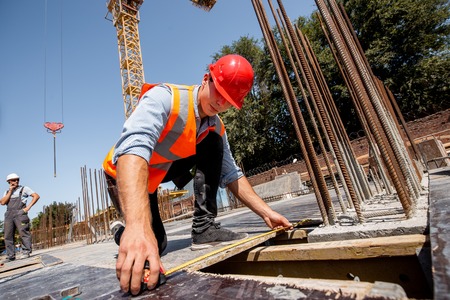 Man dressed in shirt, orange work vest and helmet measures the hole with a tape measure on the...