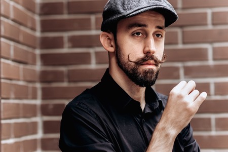 Stylish barber with mustache and beard dressed in a black shirt and flat cap stands on brick wall...