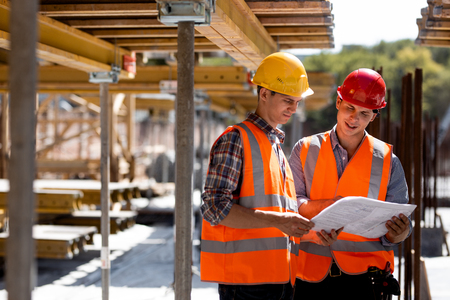 Two civil engineers dressed in orange work vests and helmets explore construction documentation on the building site near the wooden building constructions