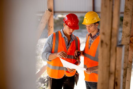 Two men dressed in shirts, orange work vests and helmets explore construction documentation on the...