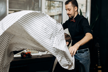 Barber dressed in a black shirt puts a cape on the stylish man sitting in the armchair in a barbershopの写真素材