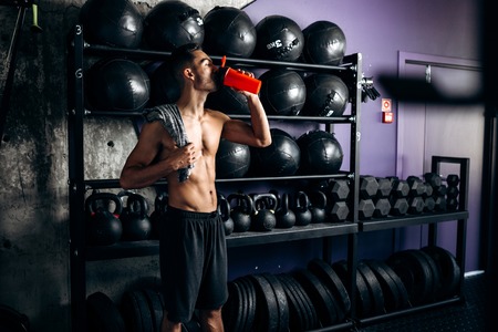 Athletic dark-haired man with a naked torso dressed in the  black shorts is standing near the sport equipment in the gym and drinking waterの写真素材