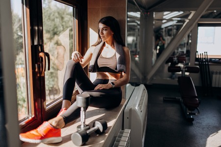 Beautiful athletic girl dressed in black sports top and tights is sitting on the windowsill with  dumbbells next to herの写真素材