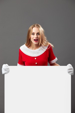 Young and beautiful mrs.Santa Claus dressed in the red robe and gloves holds  the white canvas in front of her on the gray background.の写真素材