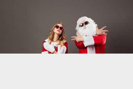 Cool gangsta Santa Claus and young beautiful mrs. Claus in sunglasses stand behind a white canvas on the gray background.の写真素材
