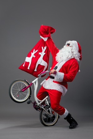 Santa Claus in sunglasses and headphones puts the bike on the back wheel and hold in the hand the bag with Christmas gifts on the white background.の写真素材