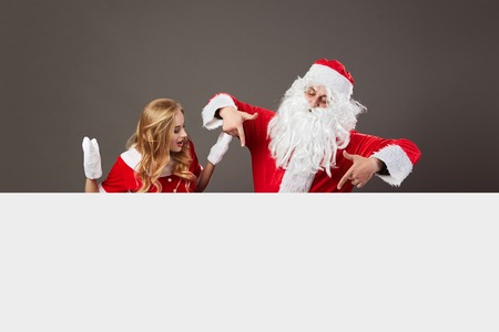 Santa Claus and young beautiful mrs. Claus stand behind a white canvas and gesticulate on the gray background.の写真素材
