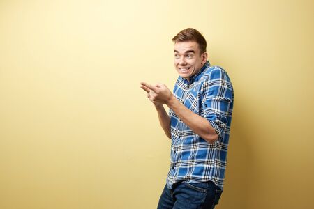 Surprised guy dressed in a plaid shirt and jeans stands next to yellow wall in the studio and points with his hands somewhereの写真素材