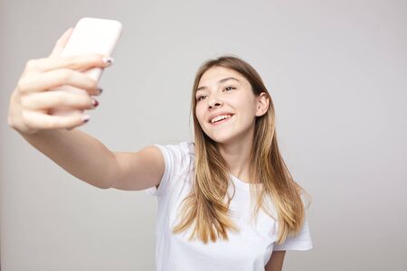Happy girl dressed in a white t-shirt makes selfie on a white background in the studioの写真素材