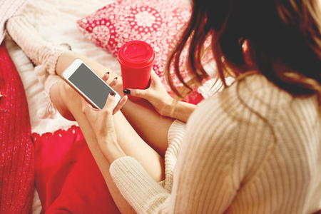 Girl dressed knitted dress and knitted socks lies on red-white blankets and pillows and holds a mobile phone and red cup of hot drinkの写真素材