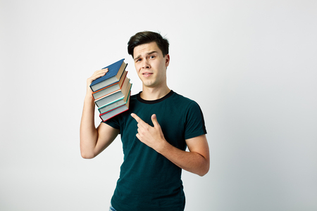 Funny dark-haired guy dressed in a dark t-shirt and jeans holds books on his shoulder on a white backgroundの写真素材