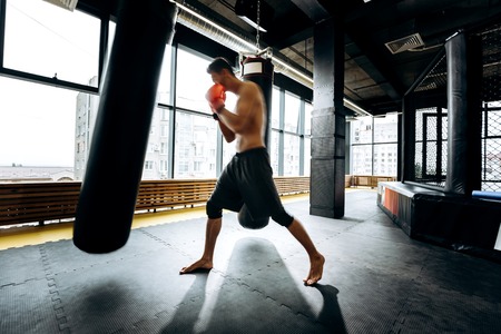 Guy in red boxing gloves with a naked torso dressed in the black shorts hits punching bag  in the...