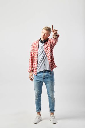 Blond guy with headphones on his neck dressed in a white t-shirt, red checkered shirt and jeans stands on the white background in the studio and point up with his fingerの写真素材