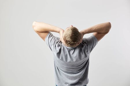 Back view of blond guy dressed in a white t-shirt that holds his hands on his head on the white background in the studioの写真素材