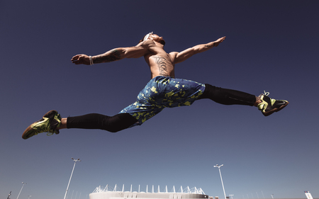 Young athletic man with a naked torso with headband dressed in the black leggings and blue shorts is jumping high in the stadium and blue sky on a warm sunny dayの写真素材