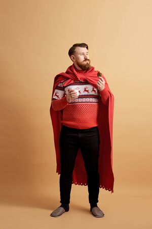 Red-haired man with beard dressed in a red and white sweater with deer and red cape  fluttering in the wind holds a glass of milk and cookie on a beige backgroundの写真素材