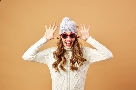 Girl dressed in white knitted sweater and hat and sunglasses is having fun on a beige background in the studioの写真素材