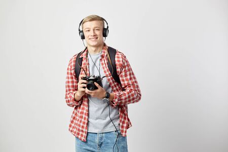 Blond guy in headphones, with black backpack on his shoulder dressed in a white t-shirt, red checkered shirt and jeans holds camera in his hands on the white backgroundの写真素材