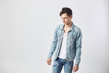 Young dark-haired guy in a white t-shirt, jeans and a denim jacket stands on the white background  in the studioの写真素材