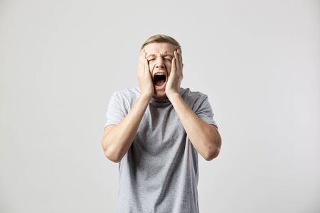 Sad guy dressed in a white t-shirt holds his hands on his cheeks and shouts on the white background in the studioの写真素材