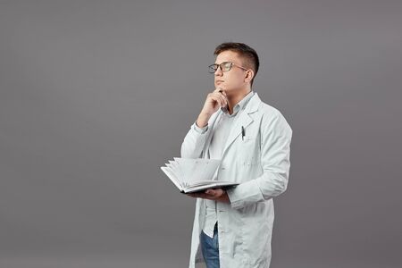 Intelligent guy in glasses dressed in a white shirt and white coat is holding a notebook and thinking on a gray backgroundの写真素材