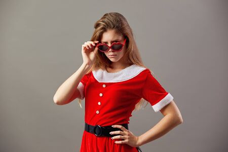 Serious charming mrs.Santa Claus in sunglasses dressed in the red robe and white gloves stands on the gray background.の写真素材