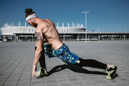 Young  athletic man with a naked torso with headband dressed in the  black leggings and blue shorts is doing stretching on paving slabs on the background of the stadium on a warm sunny dayの写真素材