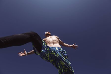 Young athletic man with a naked torso with headband dressed in the black leggings and blue shorts is jumping high on the background of blue sky on a warm sunny dayの写真素材