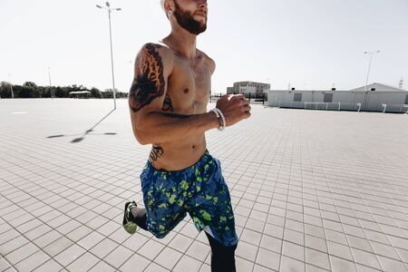 Athletic man with a naked torso with tattoos and with headband dressed in the  black leggings and blue shorts runs on paving slabs on the square on a warm sunny dayの写真素材