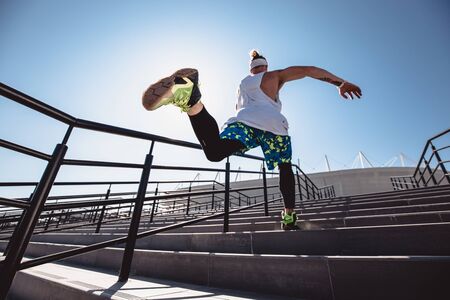 Athletic young man with headband on his head dressed in the white t-shirt, black leggings and blue shorts is jumping up the stairs outside on a sunny dayの写真素材