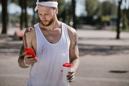 Young man with white headband dressed in a white t-shirt using mobile phone and holds a plastic cup in his hand on the sports ground outside on a sunny dayの写真素材