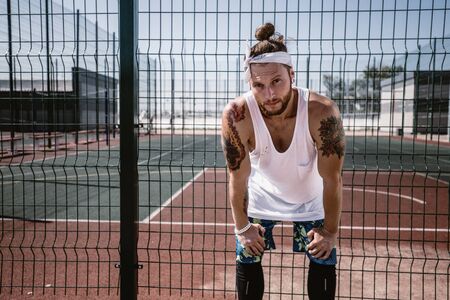 Young man with white headband  on his head and tattoos on his arms dressed in the white t-shirt, black leggings and blue shorts stands next to the playground fence outside on a sunny dayの写真素材