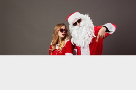 Cool Santa Claus and young beautiful mrs. Claus in sunglasses stand behind a white canvas on the gray background.の写真素材