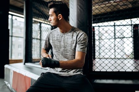 Dark-haired guy dressed in the grey t-shirt wraps a hand bandage on his hand in the boxing gymの写真素材