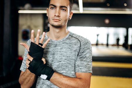 Dark-haired guy dressed in the grey t-shirt and black shorts stands  in the gym next the boxing ring and wraps a hand bandage on his handの写真素材