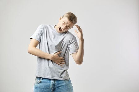 Blond guy dressed in a white t-shirt and jeans feels pain and holds hands on his stomach  on the white background in the studioの写真素材