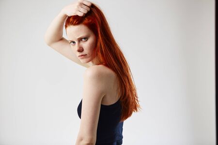 Charming redhead long-haired girl dressed in black top and jeans holds hand on her head on the white background in the studioの写真素材
