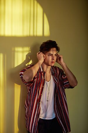 Dark-haired guy in a multi-colored striped shirt is leaning on the wall with a reflection of the sunny windowの写真素材